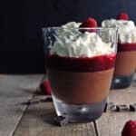 A square photo of a glass of homemade chocolate mousse topped with raspberry sauce and whipped cream.