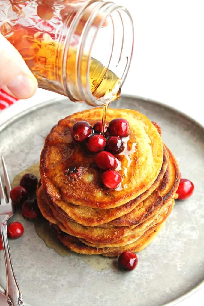 A photo of syrup pouring onto a stack on cranberry pancakes with fresh cranberries on top.