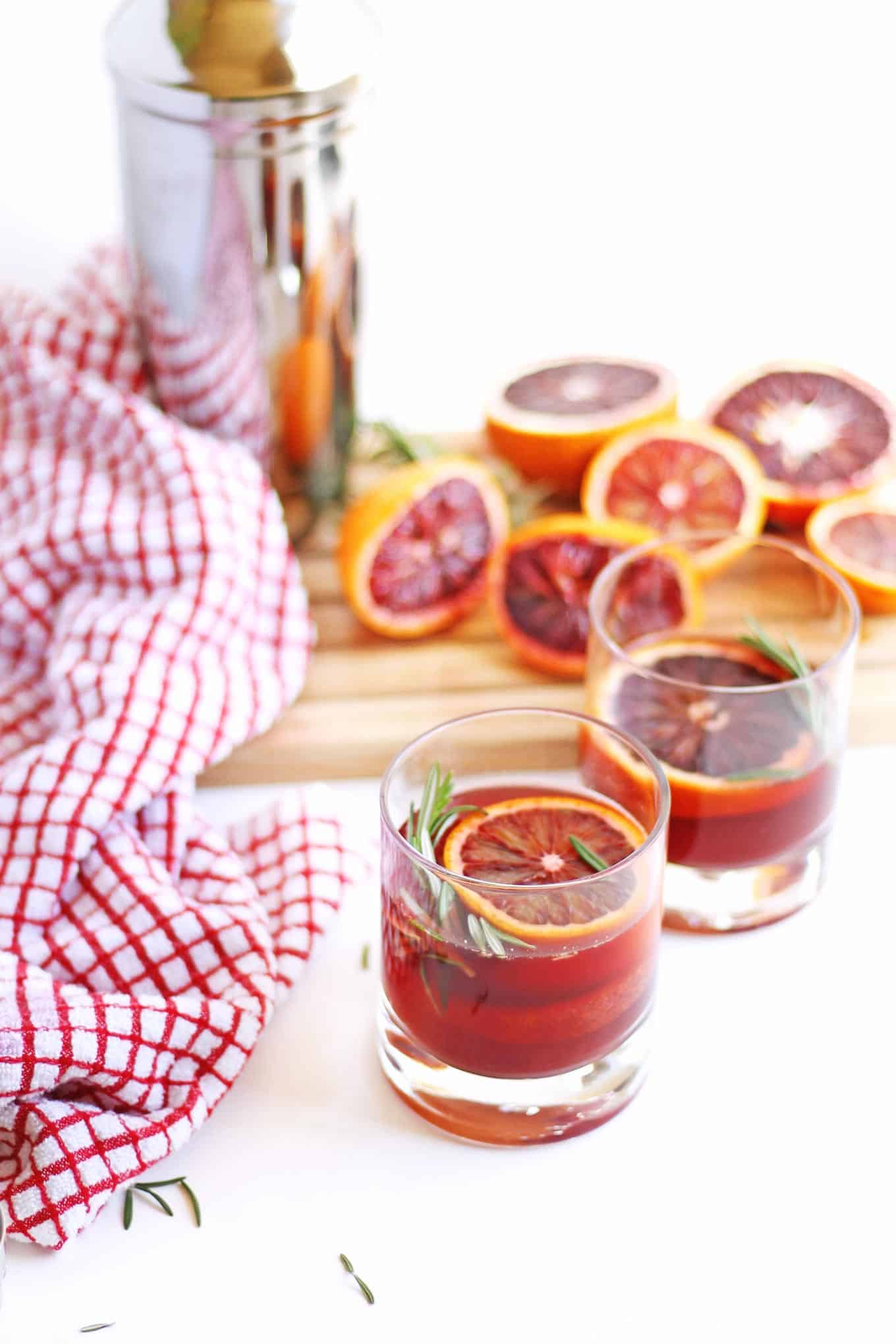 A picture of a blood orange cocktail with a sprig of rosemary and squeezed oranges in the background