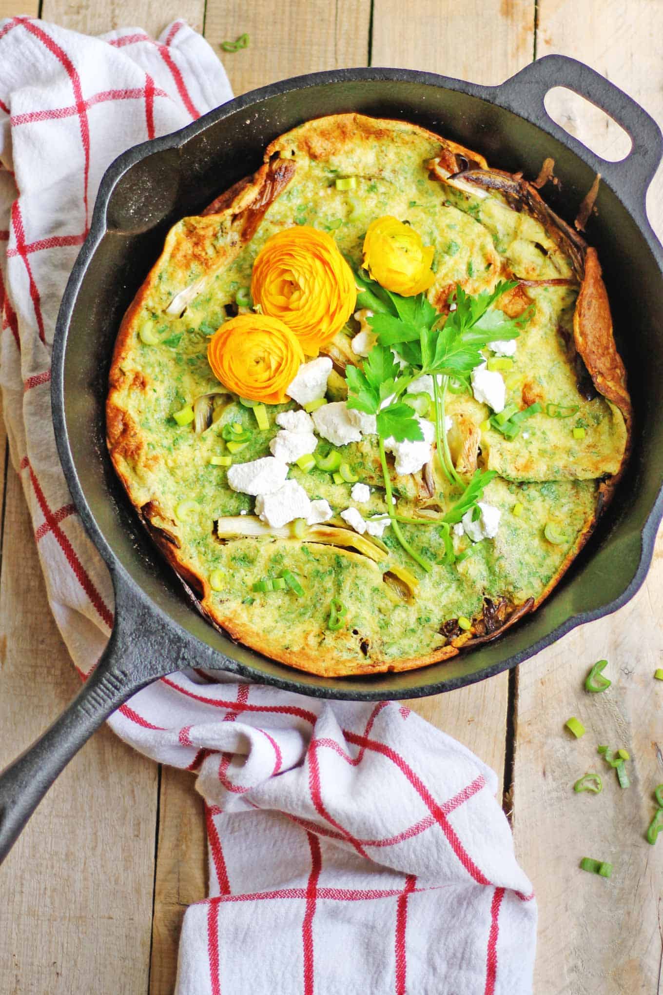 A photo of a savory dutch baby in a cast iron skillet topped with yellow flowers, parsley, and goat cheese crumbles.