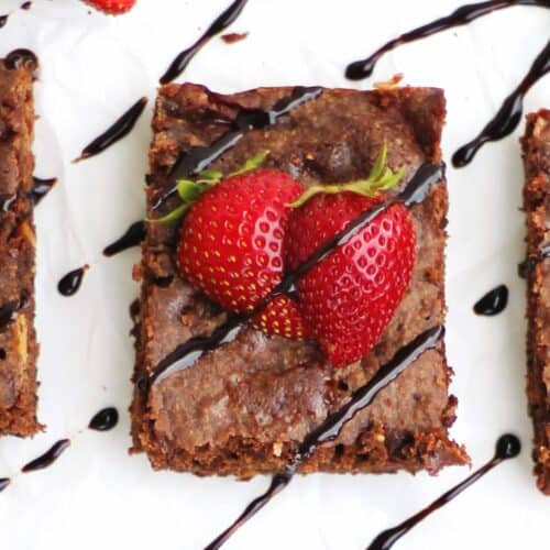 A square photo of a brownie with strawberries on top and chocolate drizzle.