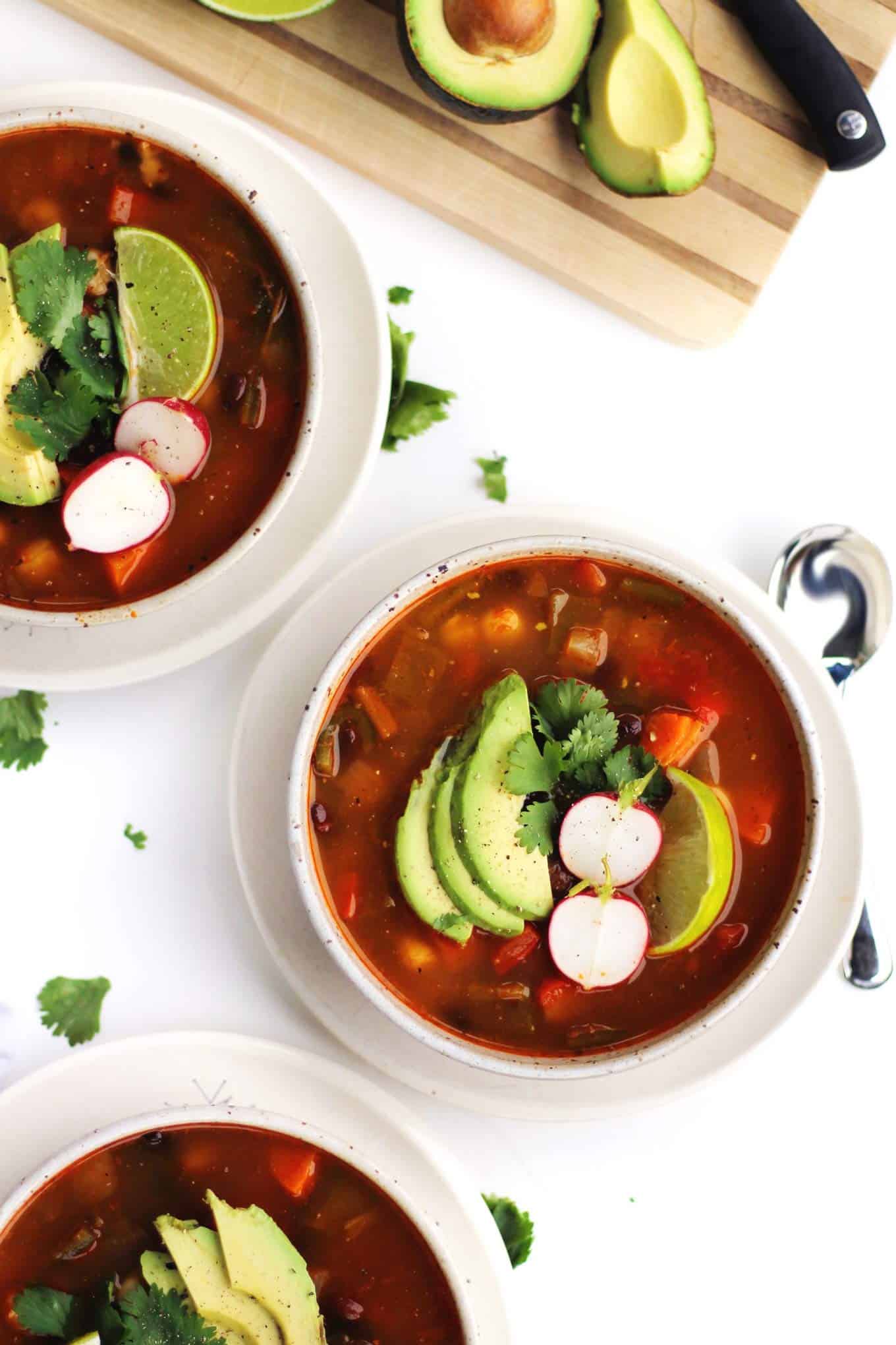 A picture of pozole in white bowls with radish, lime, cilantro, and avocado garnish.
