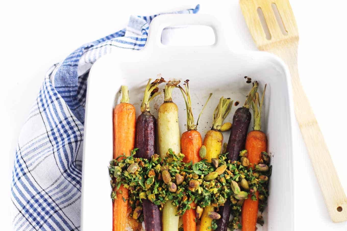 A photo of oven roasted carrots with herb relish in a white roasting pan with a blue and white plaid towel and wooden spatula.