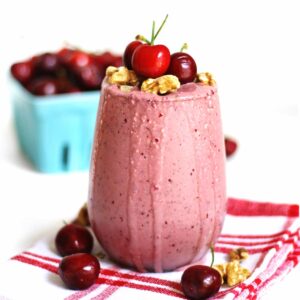 A square photo of a cherry smoothie on a red and white plaid napkin with fresh cherries and walnuts on top.