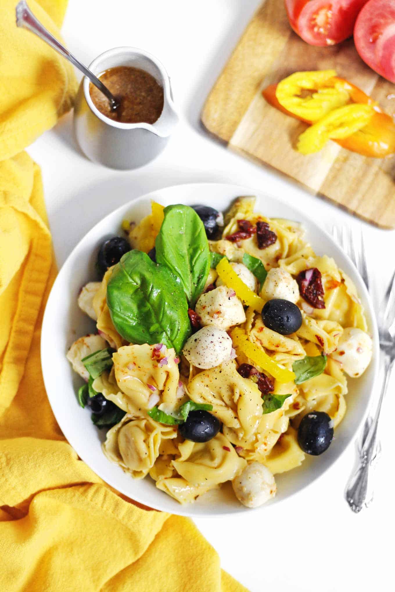 A picture of Italian tortellini salad with basil leaves in a white bowl next to a yellow cloth napkin.