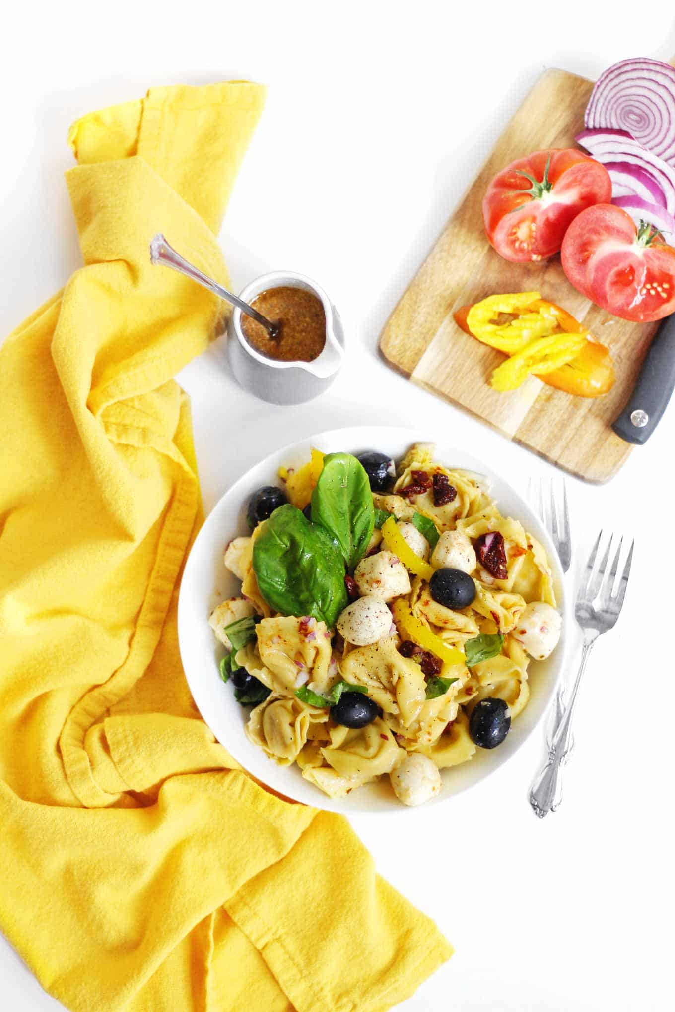 A picture of a bowl of vegetarian Italian tortellini pasta salad next to two forks and a yellow cloth napkin.