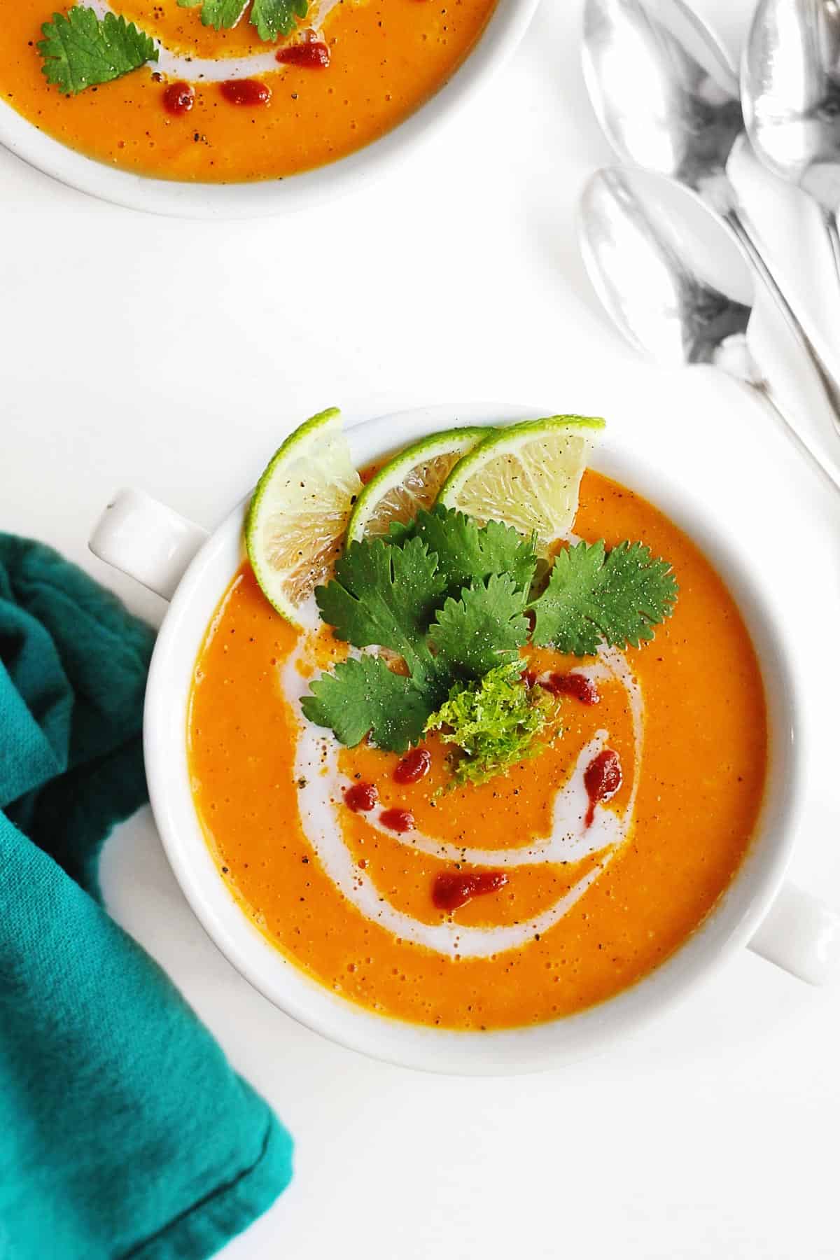A photo of pureed Thai curried butternut squash soup in a white soup bowl topped with coconut milk swirl, Sriracha sauce, lime zest and slices, and cilantro.