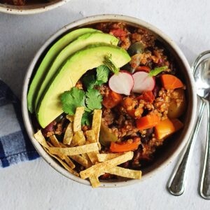 A square photo of vegan quinoa chili with tortilla strips, avocado, cilantro, and radish.