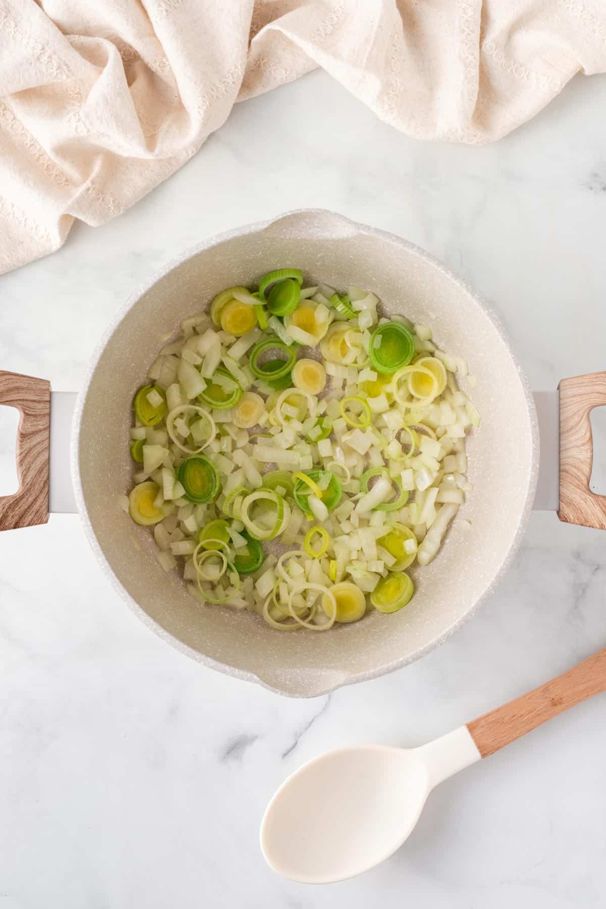 A picture of cooking onions and leeks in a oil in a pan with wooden handles.