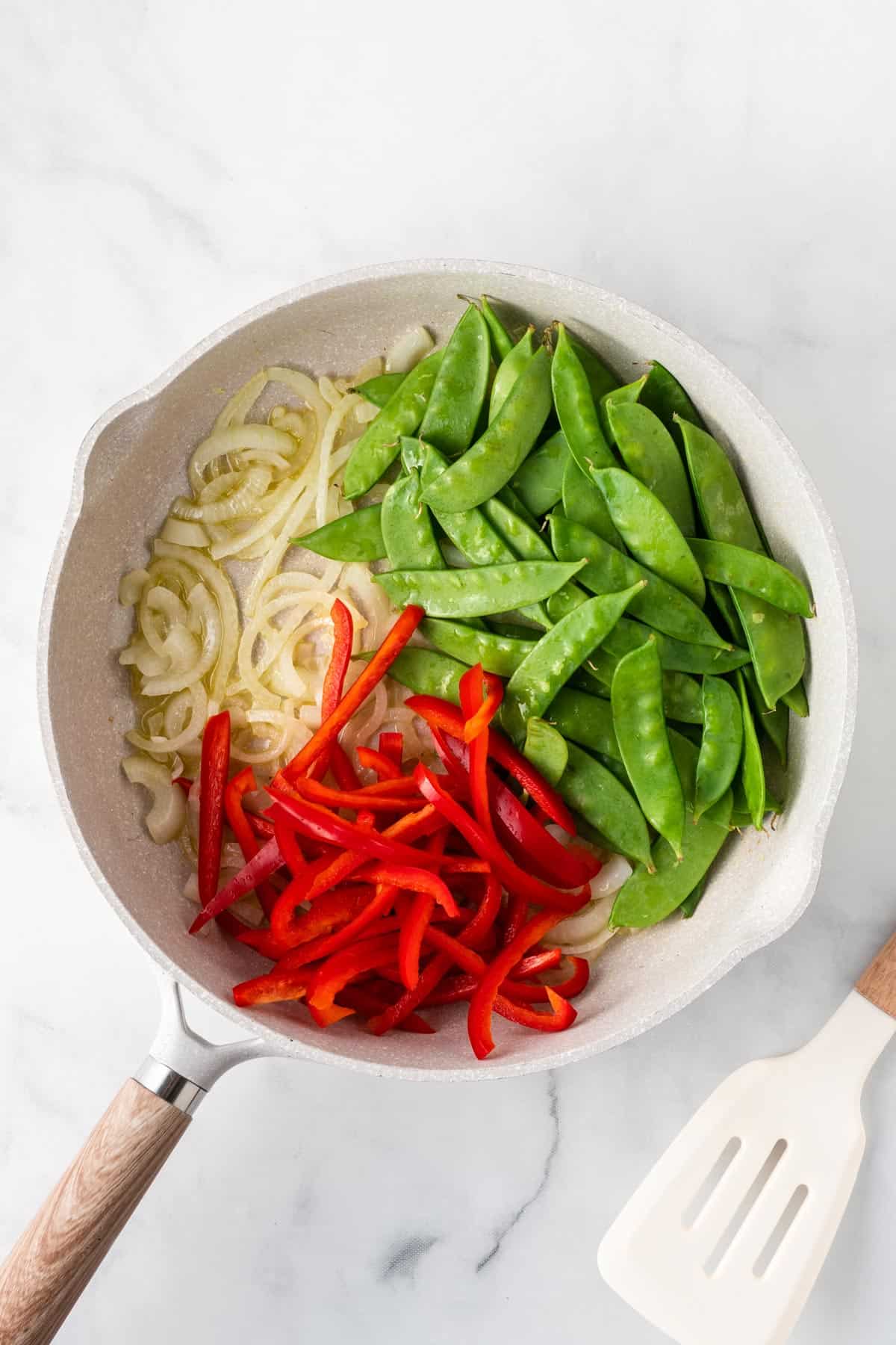 A picture of onions, snap peas, and red pepper slices in a white skillet.