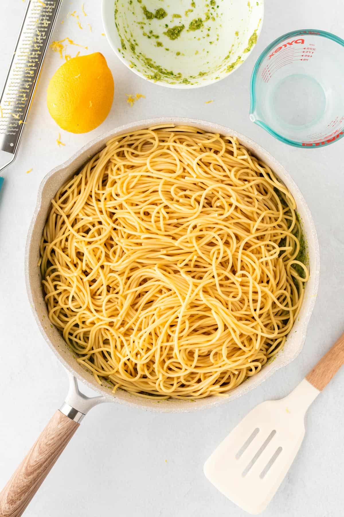 A photo of spaghetti noodles in a white skillet surrounded by a lemon and a spatula.