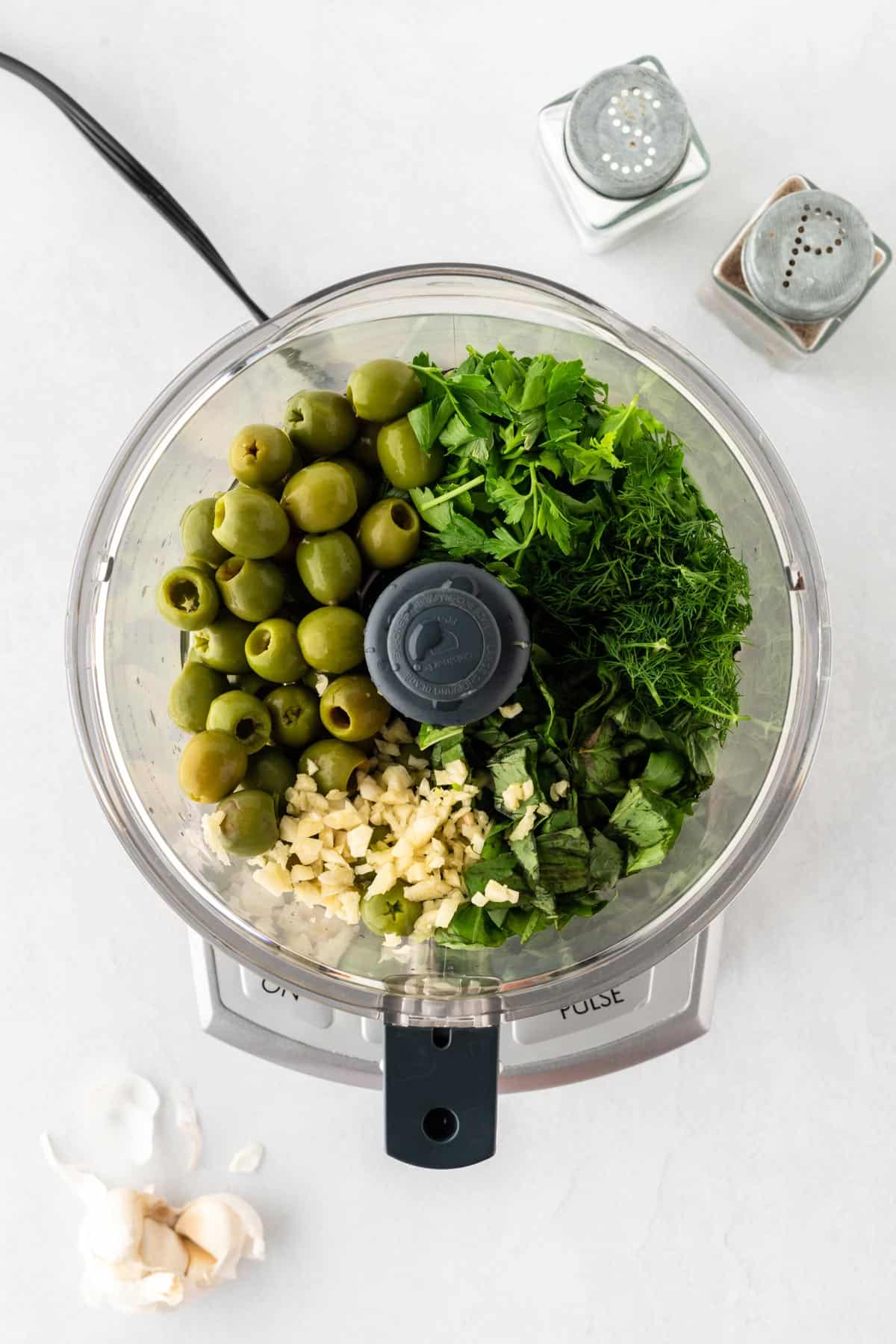 A photo of green olives, herbs, and garlic in a food processor.