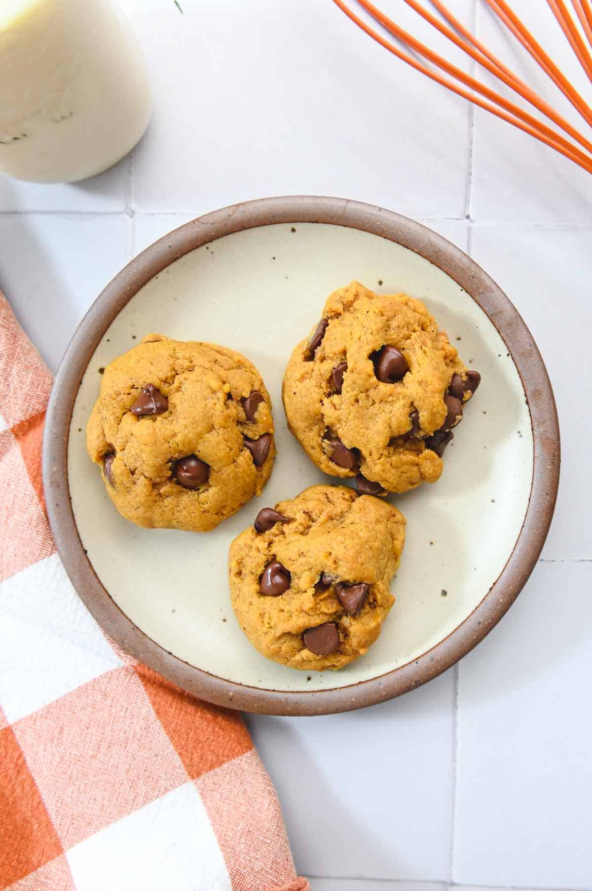 A photo of 3 pumpkin chocolate chip cookies on a brown plate.