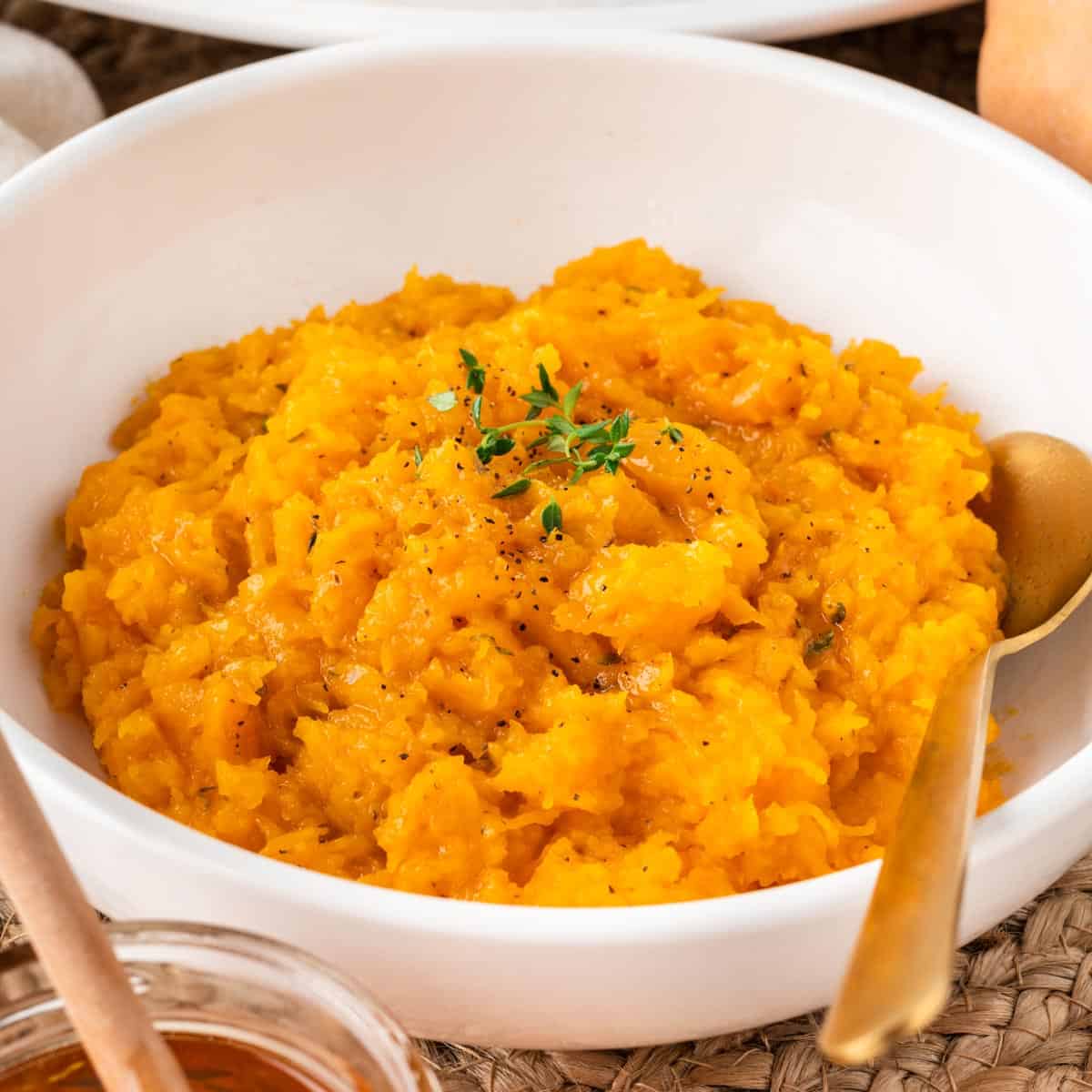 A square photo of mashed butternut squash in a white bowl with a gold spoon.