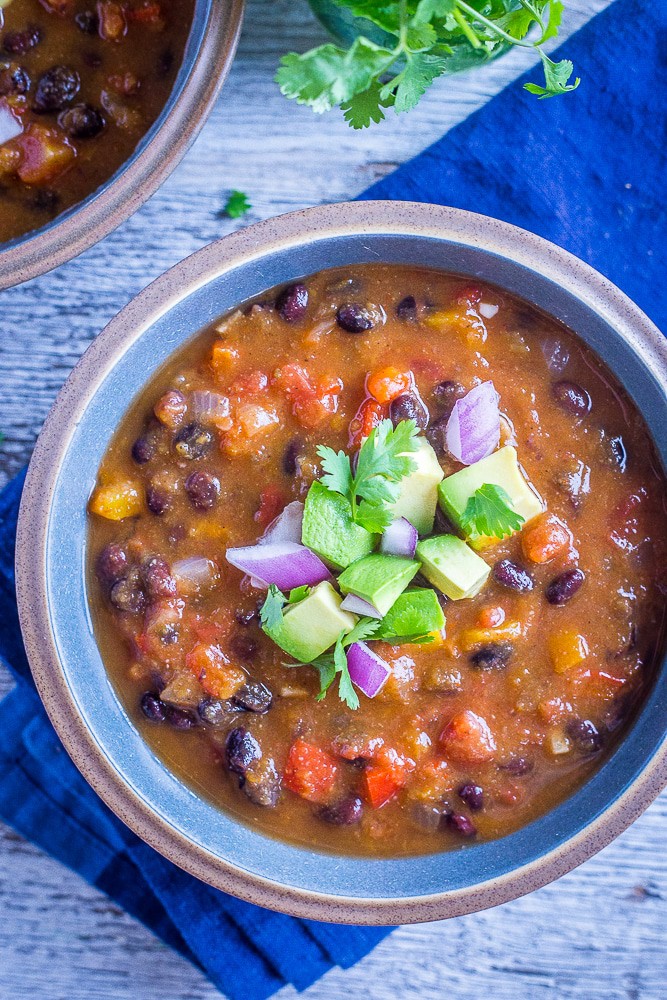 A photo of a bowl of chili topped with avocado, red onion, and cilantro.