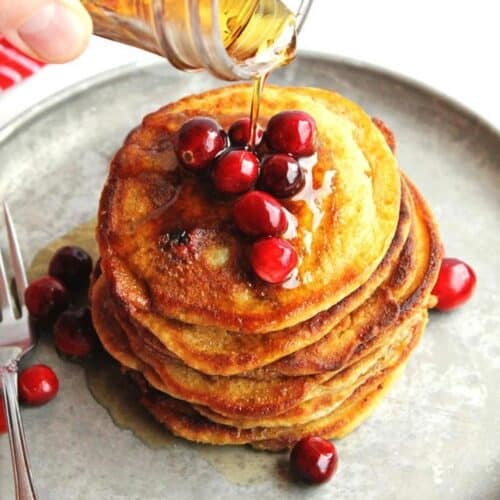 A square photo of pouring syrup on a stack of pancakes with fresh cranberries on top.