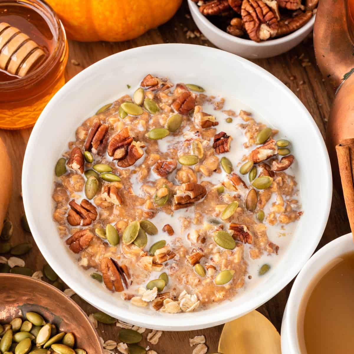 A square photo of pumpkin oatmeal in a white bowl with pecans and pepitas on top.
