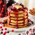A square photo of a stack of cranberry pancakes with cranberry sauce, butter, and sugared cranberries on top.