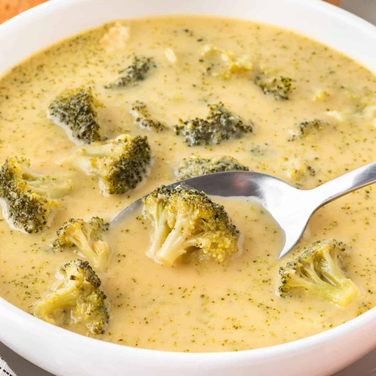 A square photo of a spoon scooping up broccoli out of a bowl of broccoli soup.