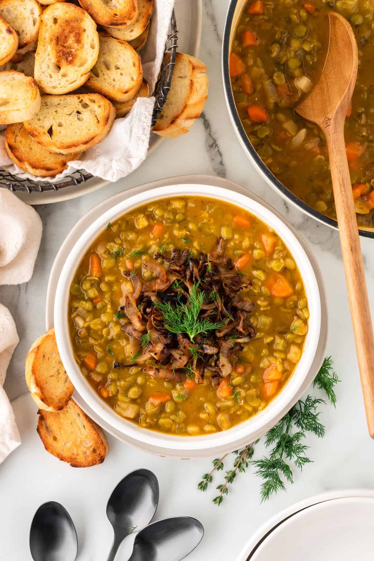 A photo of a bowl of vegetarian split pea soup with bread slices and spoons next to it.