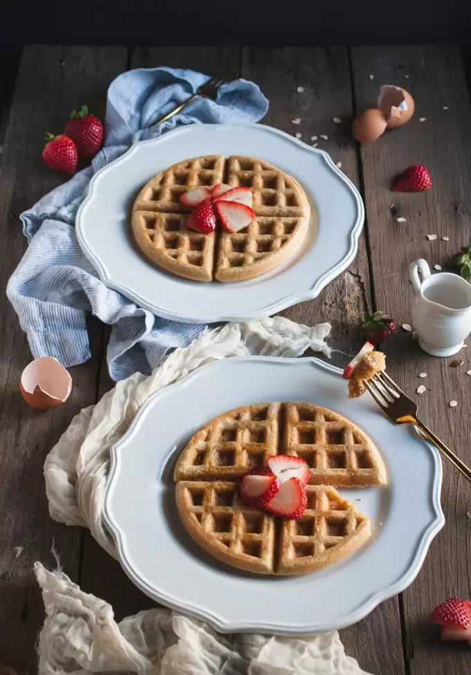 A photo of two white plates with waffles topped with strawberries on them.