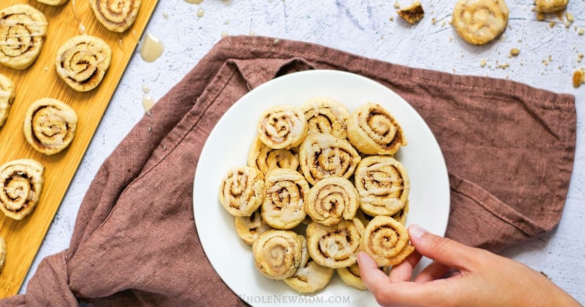A photo of a hand grabbing a cinnamon roll cookie from a plate of them.