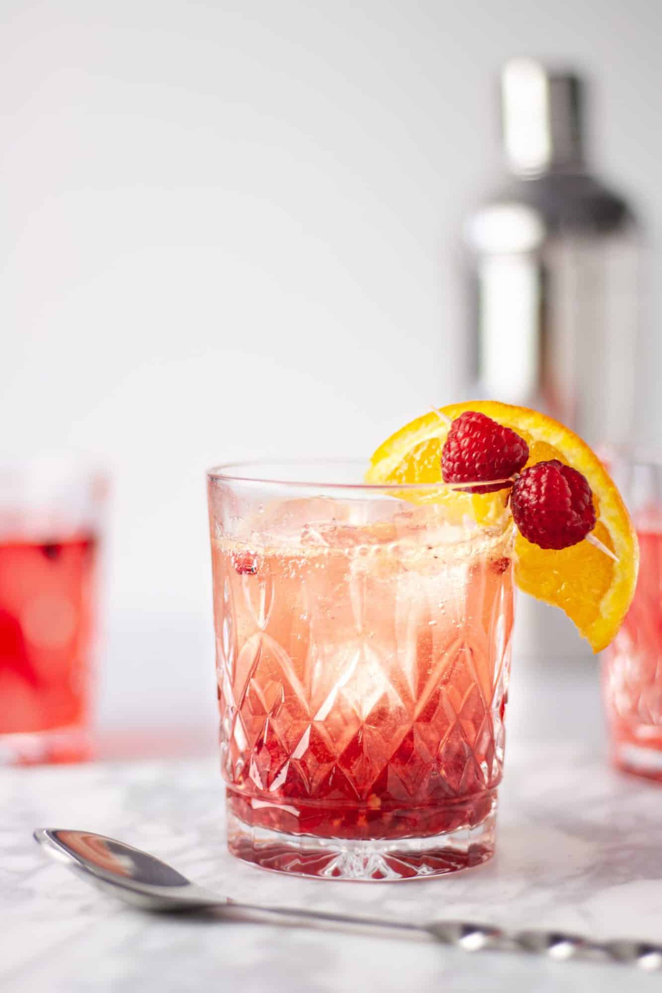 A photo of a bourbon raspberry cocktail with fresh raspberry and orange garnish.