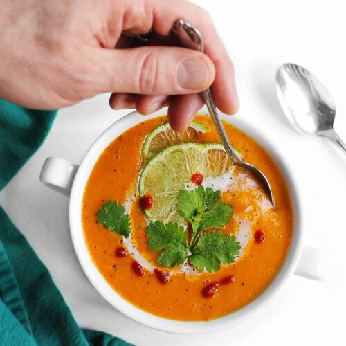 A square photo of dipping a spoon into a bowl of butternut squash soup with thai butternut squash soup toppings.
