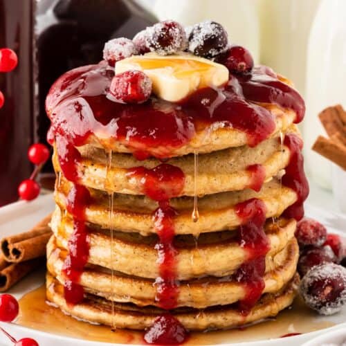 A square photo of a stack of pancakes with cranberry sauce drizzled on top.
