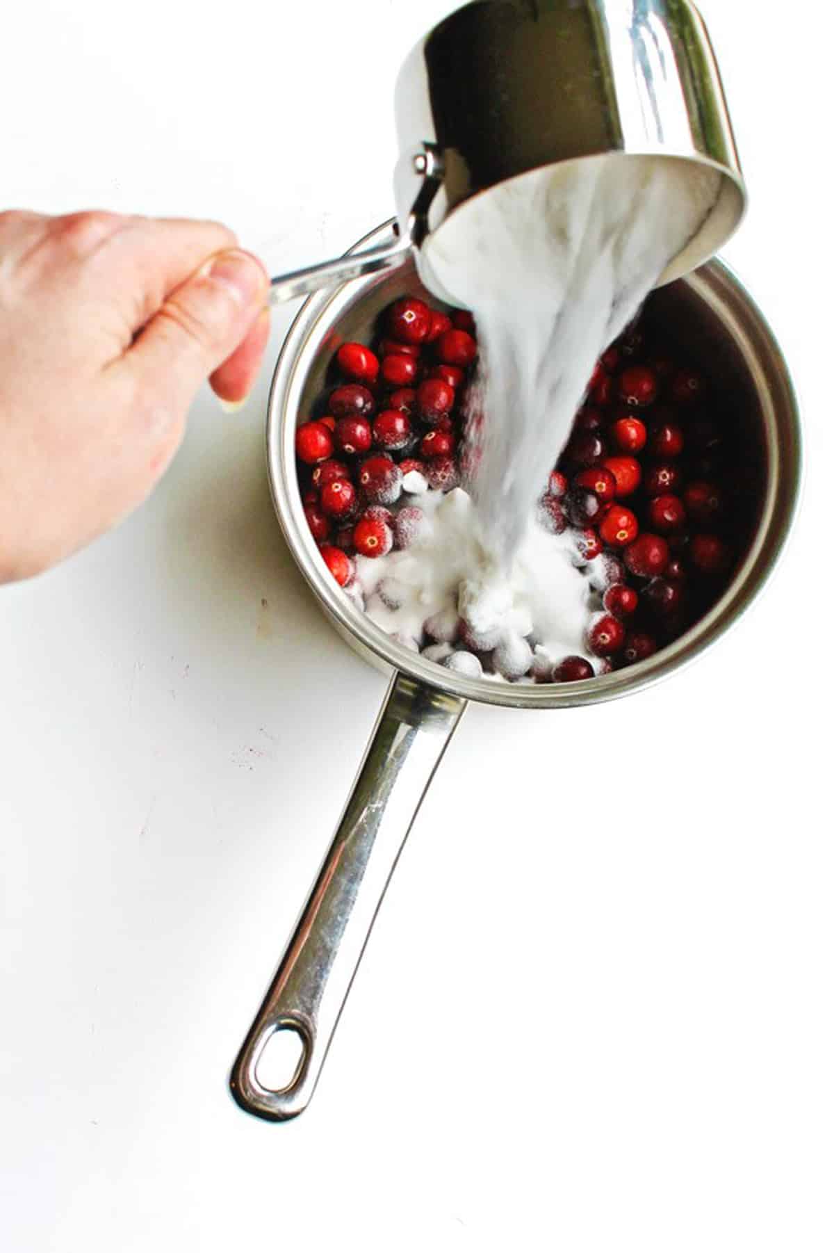 A photo of pouring sugar into a sauce pan with cranberries.