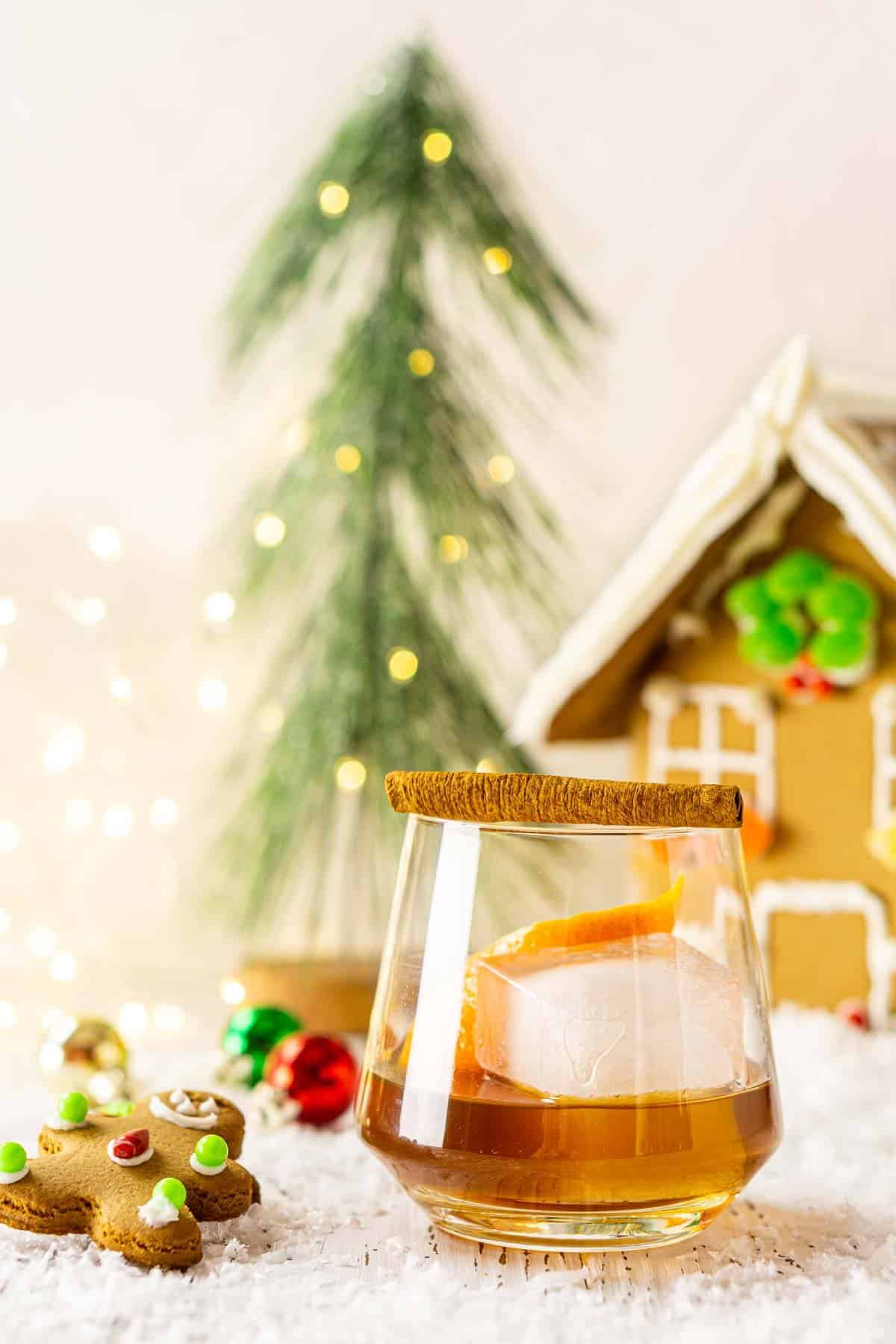 A photo of a gingerbread old fashioned cocktail with a gingerbread house in the background.